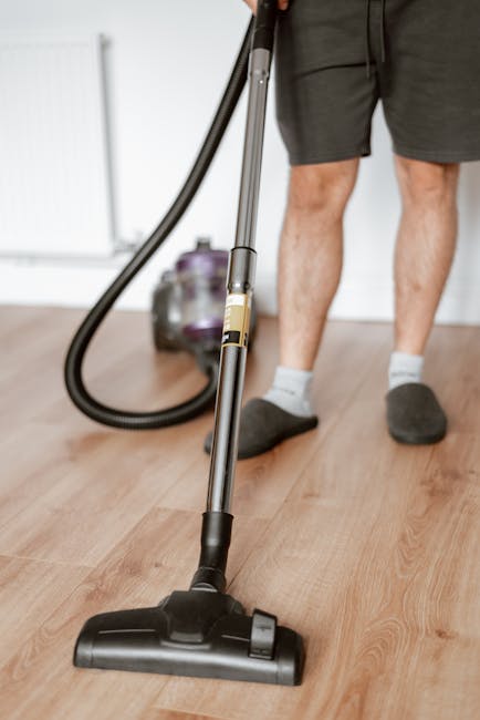 Close-up view of a person vacuuming a light wood laminate floor in a residential living room, with visible vacuum cleaner hose and metal wand. The individual is wearing dark shorts, white socks, and slippers, and is operating a modern upright vacuum cleaner to perform surface cleaning and dust removal. In the background, a purple and black vacuum canister is partially visible, along with a white wall and minimal furniture, highlighting a clean and well-maintained space. This scene exemplifies professional domestic cleaning, emphasizing hygiene and deep cleaning processes, as offered by Balham Carpet Cleaning near Balham Station SW12.