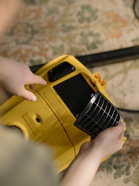 A person operating a yellow industrial carpet cleaning machine, holding the handle with one hand and adjusting the black control panel with the other, situated on a patterned area rug with warm tones in a residential setting. The machine’s front vent is visible, indicating a deep cleaning process. The surrounding area features a wooden floor and possibly a wall or furniture with muted colors, demonstrating effective surface cleaning and maintenance by Balham Carpet Cleaning, as referenced on the Balham carpet cleaning near Balham Station SW12 webpage.