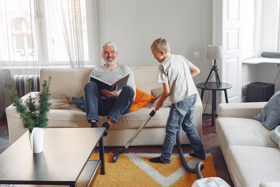 A young boy vacuuming a beige sofa in a well-lit living room with white walls and large windows, while an older man with gray hair and a beard sits on the sofa reading a book, surrounded by a black round side table, a lamp, and houseplants, demonstrating domestic surface cleaning and maintenance with a focus on cleanliness and hygiene, within a tidy, modern home environment. The scene is associated with Balham Carpet Cleaning's urgent same-day sofa cleaning services on Bedford Hill.