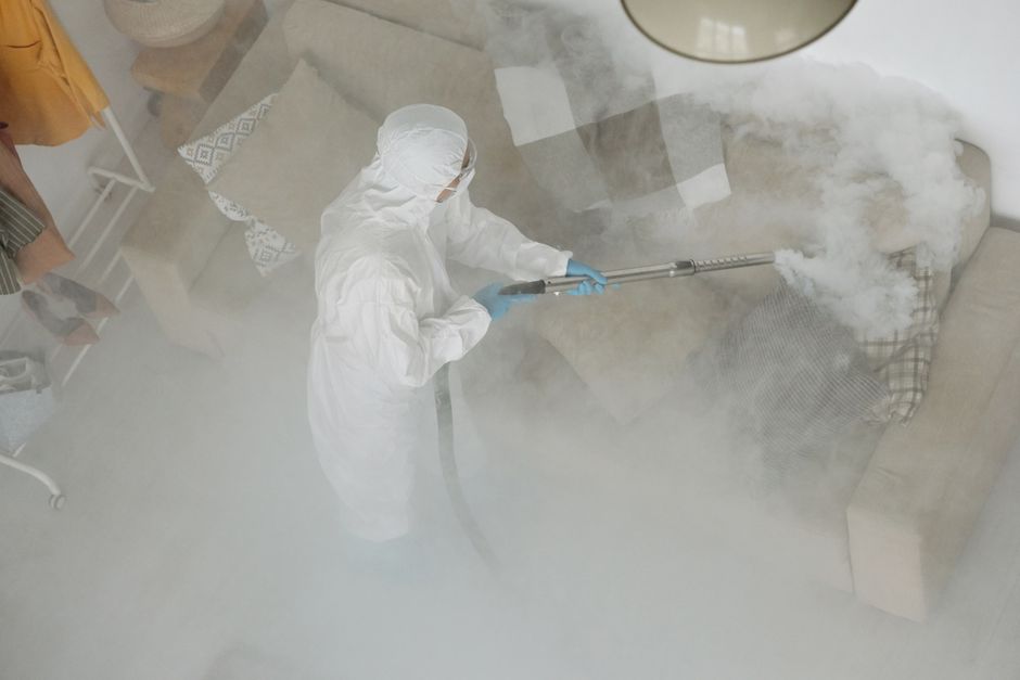 A professional cleaner dressed in a white protective suit and blue gloves is performing surface cleaning with a steam or fogging device, emitting dense white vapor across a beige fabric sofa in a well-lit living room. The room features a wooden side table, a patterned rug, and a light-colored floor. Visible surfaces include the sofa's upholstery, the floor, and parts of the surrounding furniture, all appearing clean and dust-free. The image emphasizes deep cleaning and sanitisation processes carried out by Balham Carpet Cleaning at Bedford Hill with a focus on maintaining hygiene in a domestic setting.