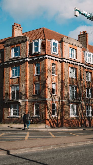 The image shows a multi-story brick residential building with pitched red-tiled roofs, large sash windows, and chimney stacks, situated along a city street under a partly cloudy sky. Shadows of leafless trees fall onto the facade, casting intricate patterns across the brickwork. Two pedestrians walk on the sidewalk in front of the building, which is bordered by a curb and road markings. A construction crane is visible in the background, indicating ongoing urban development. The scene highlights an urban setting where professional cleaning services by Balham Carpet Cleaning could be performed to maintain the property's exterior and surrounding areas, aligning with the service category of surface cleaning and deep cleaning for residential or commercial properties as detailed in the 'End of tenancy cleaning Balham Broadway flats SW12, BALHAM' page.