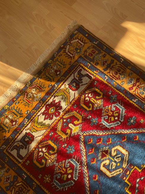 Close-up view of a colorful, patterned rug with intricate geometric and floral motifs, partially rolled back to reveal a light wooden floor beneath. The rug features shades of red, blue, yellow, and cream, with fringed edges along one side. The surface appears clean and well-maintained, reflecting good cleaning and restoration. Sunlight from a nearby window illuminates the scene, highlighting the texture and vibrant colors of the rug. This image exemplifies surface cleaning and deep cleaning techniques that Balham Carpet Cleaning offers for domestic and commercial spaces, ensuring hygiene and preservation of valuable textiles in a residential setting.
