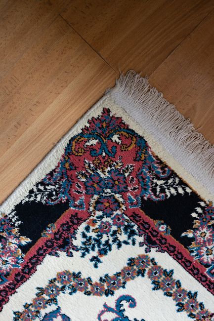 Close-up image of a patterned rug with intricate floral and scroll motifs in shades of red, blue, and black on a cream background, placed on a wooden floor in a room with ample natural light. The rug features fringed edges in white, and the surface appears clean and well-maintained, highlighting the surface cleaning and deep cleaning services offered by Balham Carpet Cleaning as part of their domestic cleaning and sanitisation solutions for residential spaces in the Nightingale Triangle area.