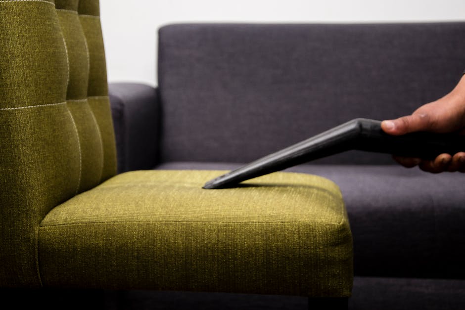 A close-up view of a hand holding a vacuum cleaner attachment, thoroughly cleaning the olive-green upholstered seat cushion of a modern armchair in a living room setting. The chair features a tufted backrest with visible stitching, and the fabric surface appears clean and well-maintained. In the background, a dark grey sofa with a smooth fabric finish is partially visible, arranged against a plain white wall. Bright, natural lighting highlights the fabric textures and the cleanliness of the furniture, indicative of professional surface cleaning aimed at maintaining hygiene and freshness in a domestic environment. The scene emphasizes detailed deep cleaning practices, consistent with the services offered by Balham Carpet Cleaning, as referenced on the Tooting Bec Lido rugs and upholstery cleaning page.