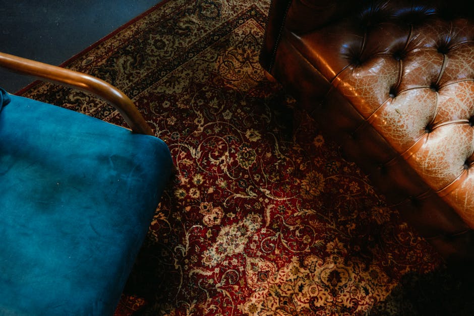 Close-up of a deep red ornate patterned rug with intricate floral and scroll motifs, situated on a wooden floor. Part of a vintage brown leather Chesterfield sofa with tufted upholstery is visible in the top right corner, showing signs of wear and some discoloration. To the left, a portion of a modern blue upholstered chair with a smooth fabric surface and wooden armrest is seen, contrasting with the traditional rug and sofa. The room is softly lit, highlighting the detailed design of the rug and the textures of the furniture surfaces, emphasizing the importance of surface cleaning and maintenance in domestic spaces, as offered by Balham Carpet Cleaning.
